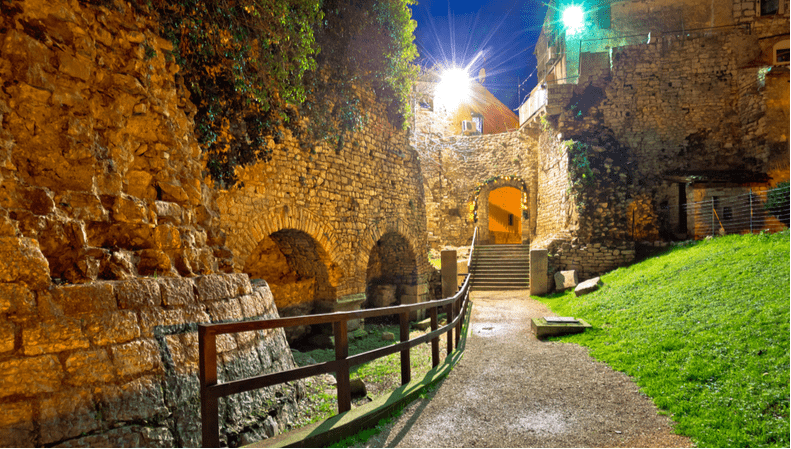 Illuminated Poreč city walls at night, featuring stone arches and a pathway
