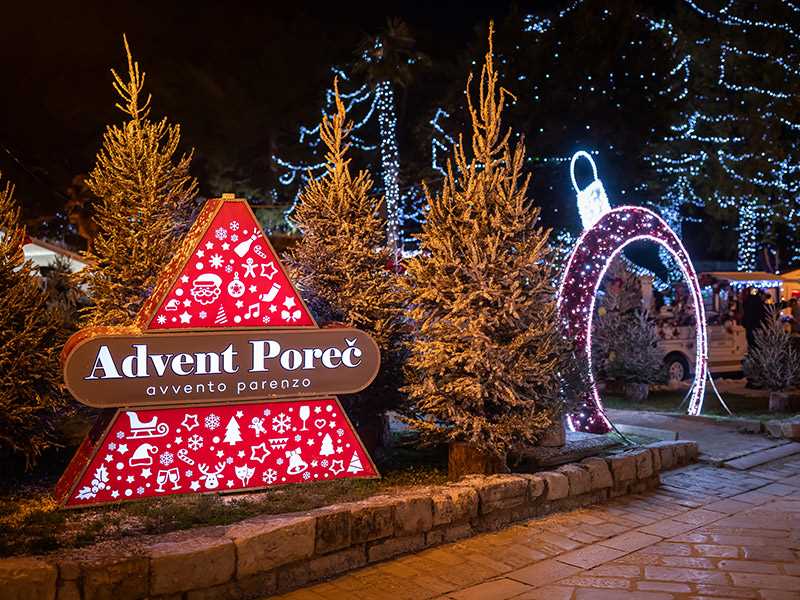 Ein festliches Weihnachtsmarktschild, umgeben von geschm&uuml;ckten B&auml;umen und glitzernden Lichtern in Poreč