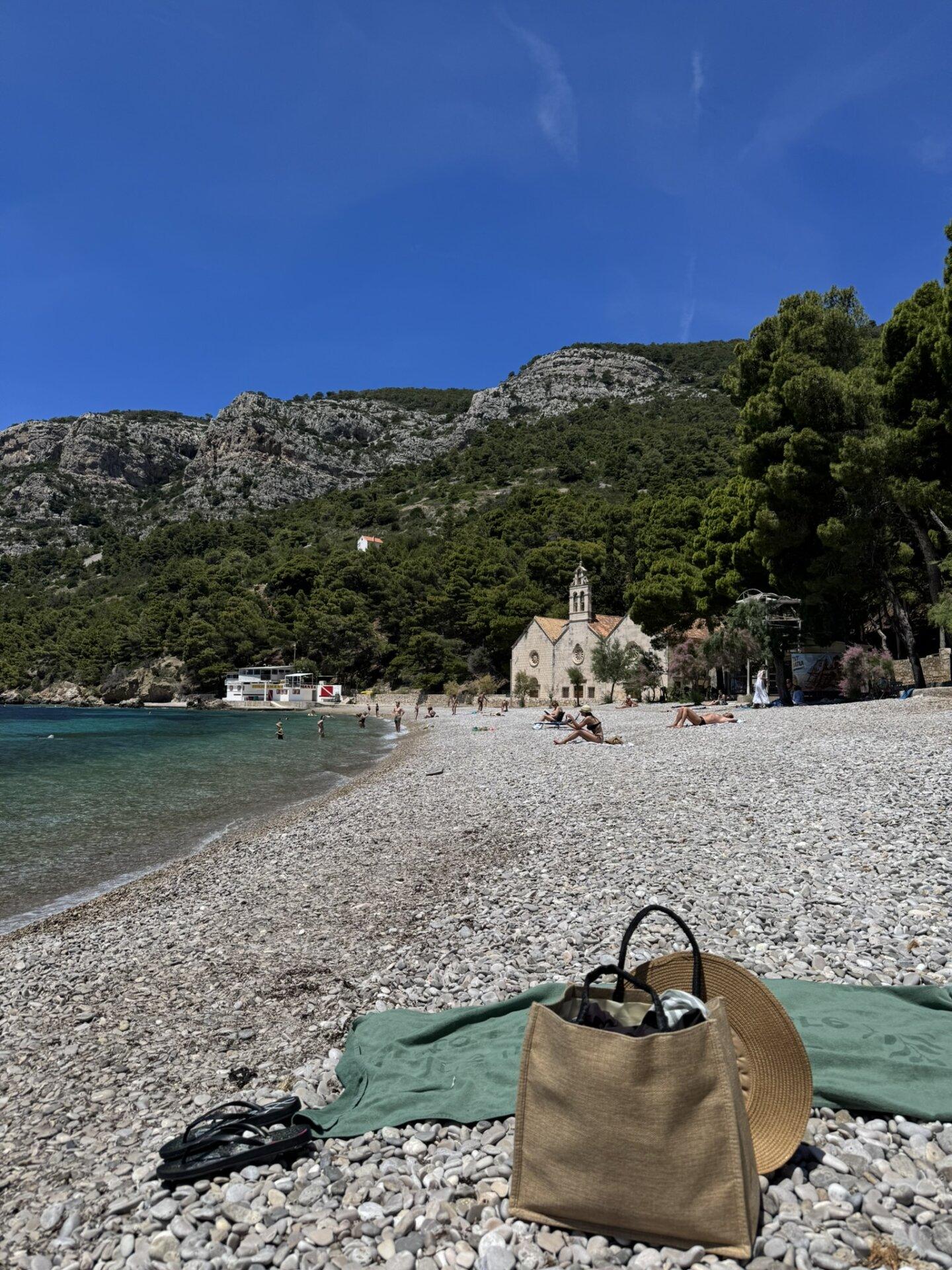 Šljunčana plaža u Komiži na otoku Visu, s tirkiznim morem, planinama u pozadini, te torbom i šeširom na zelenom ručniku