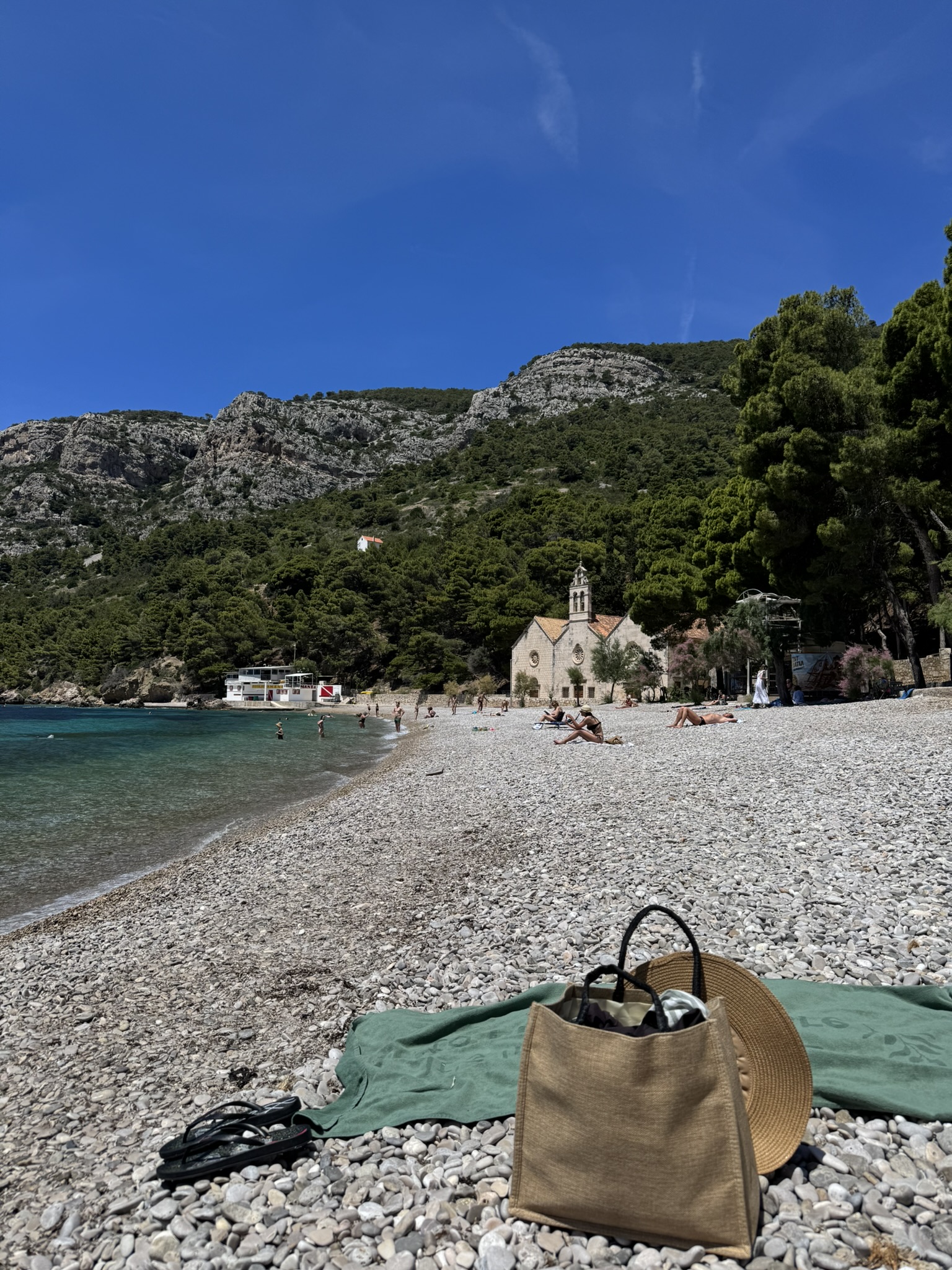Pebble beach in Komiža, Vis island, with turquoise water, mountains in the background, and a beach bag and hat on a green towel