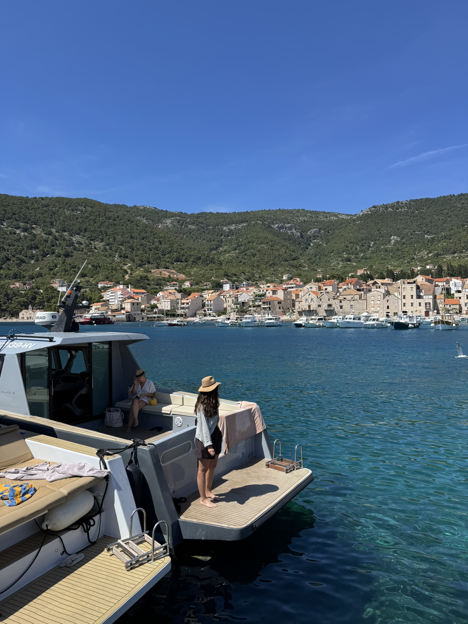 Woman standing barefoot on the back of a boat, looking toward Komiža village on Vis island with green hills in the background