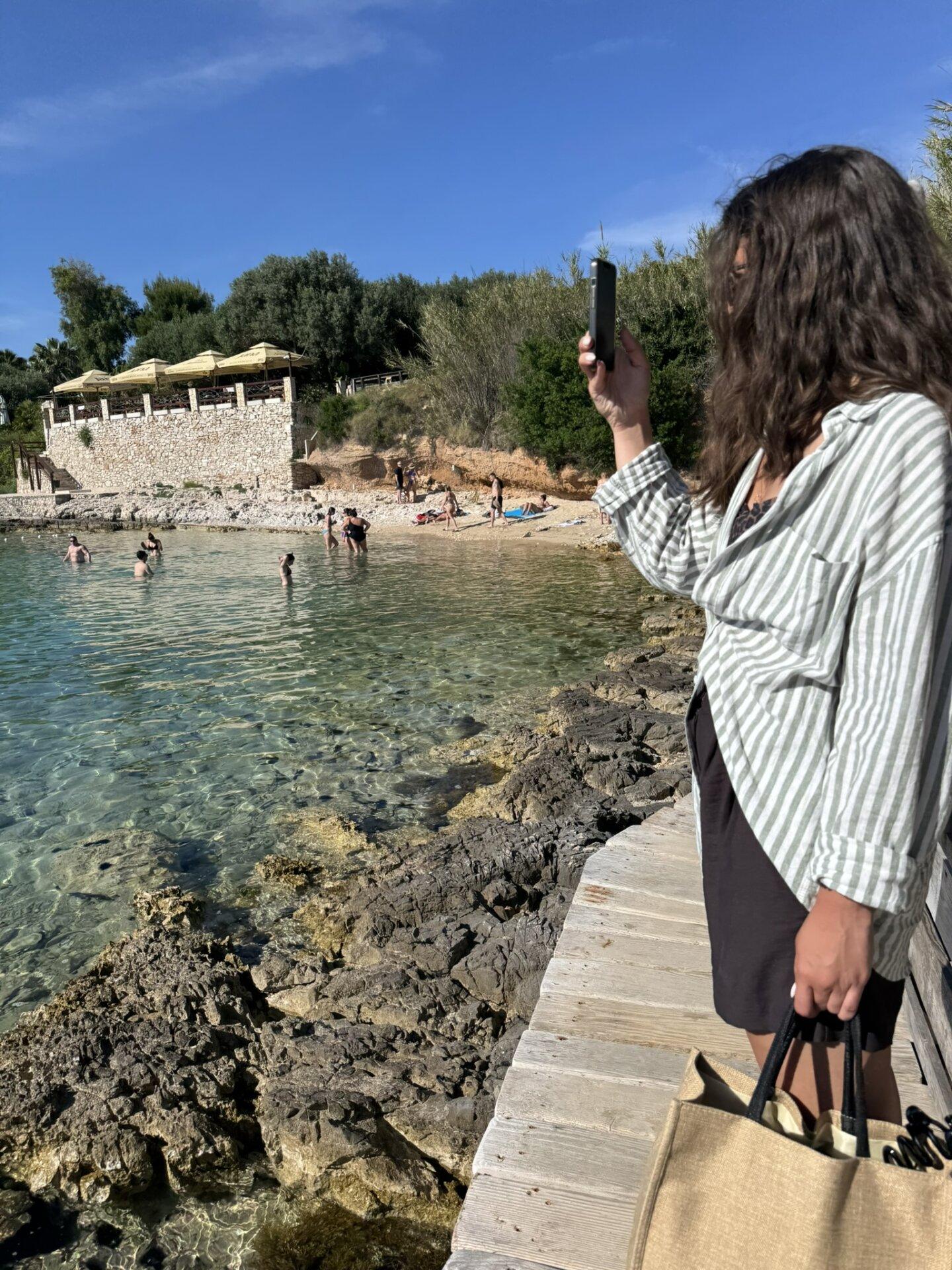 Žena stoji na drvenoj stazi uz kamenitu obalu na plaži Palmižana, fotografira plivače i prizor s plaže.