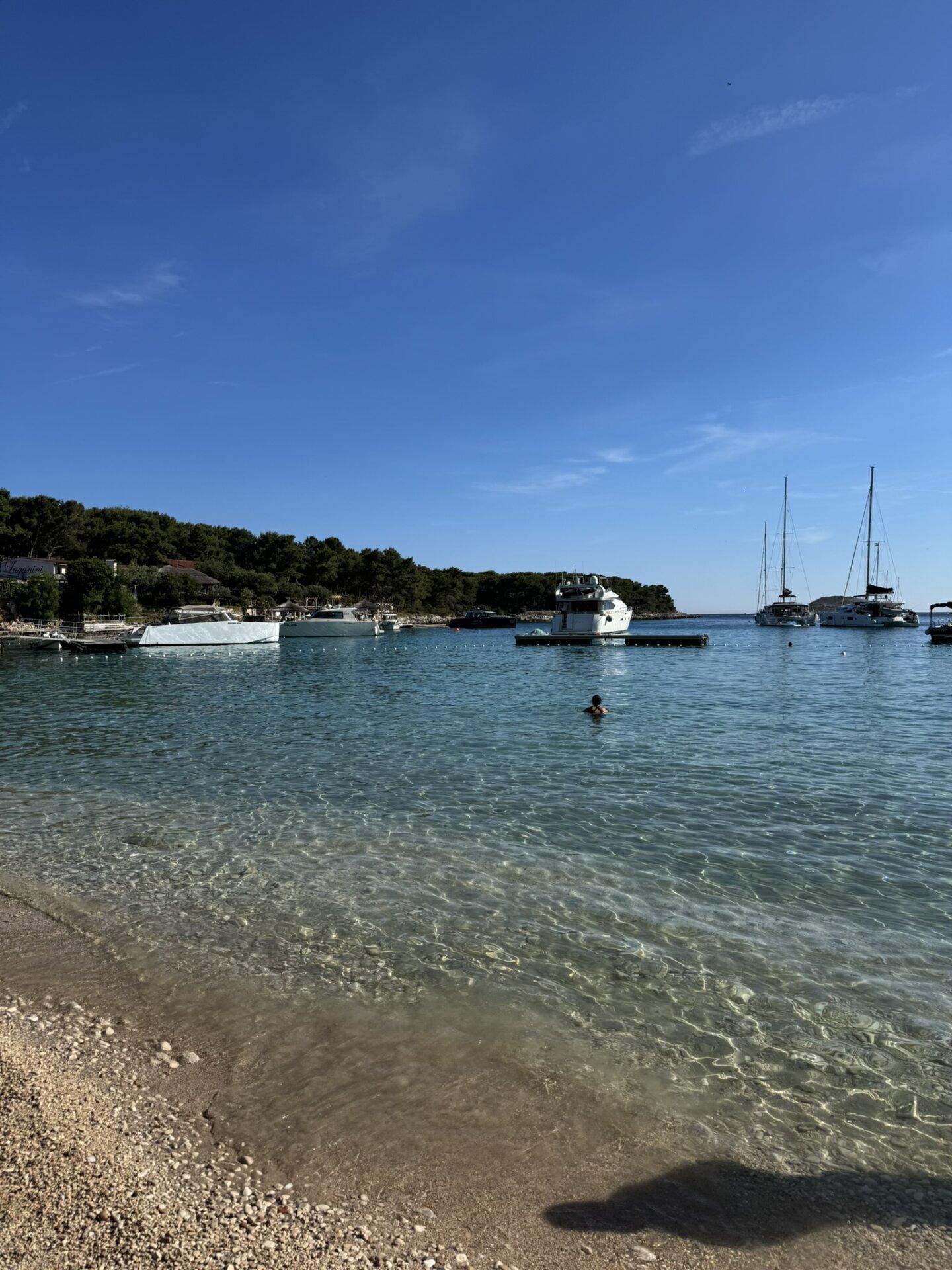 Pogled s obale plaže Palmižana, s bistrom tirkiznom vodom, usidrenim brodovima i plivačem u moru.
