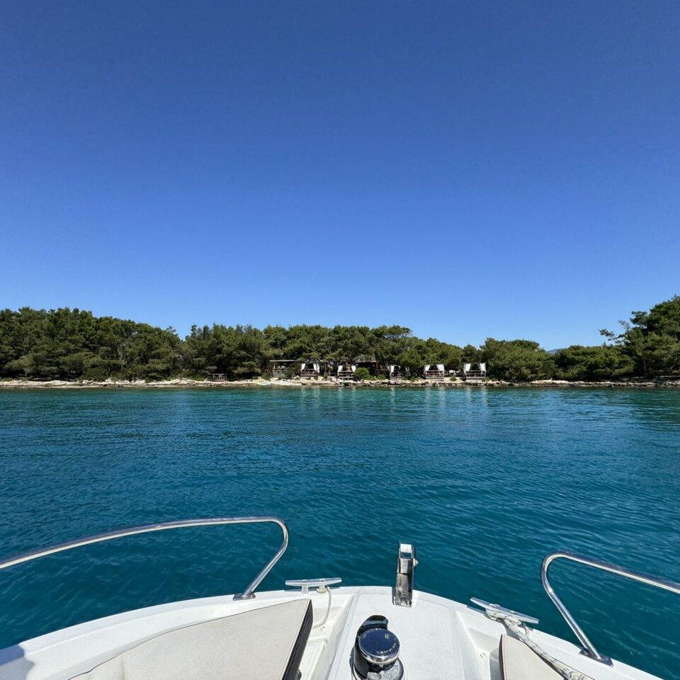View from a boat approaching a secluded pine-covered island with rustic cabanas along the shoreline and clear turquoise water