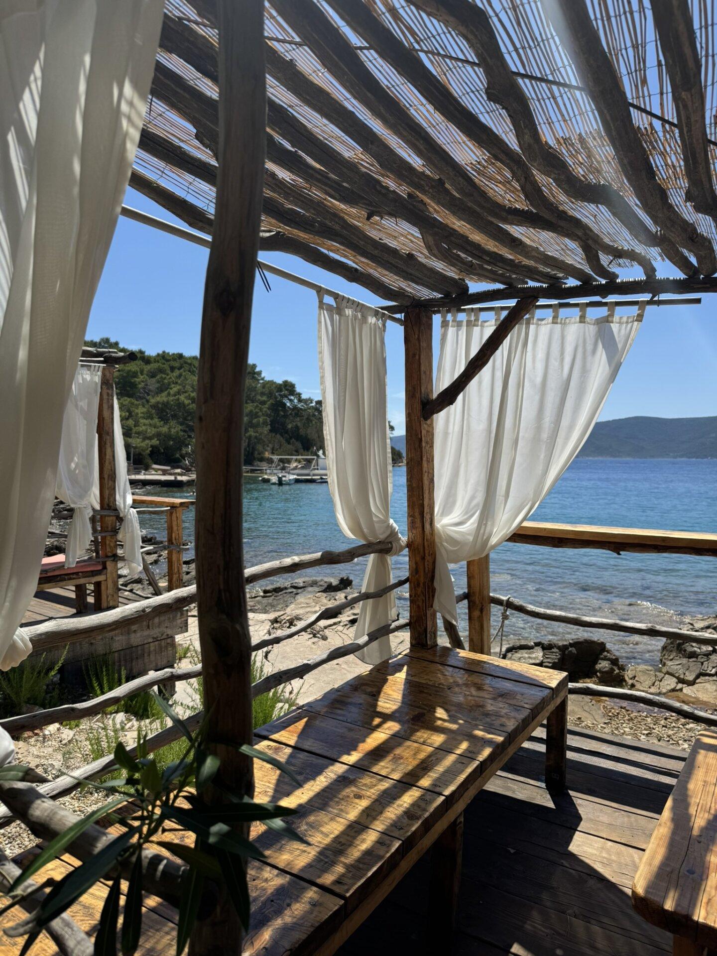 Rustikalna drvena kabana s bijelim zavjesama s pogledom na kamenitu plažu i bistro plavo more, u hladu pod krovom od naplavljenog drva.