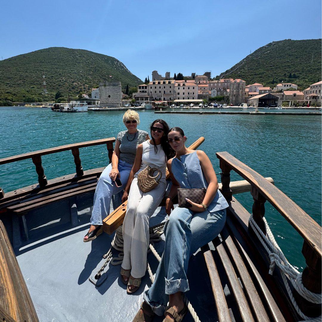 Drei Frauen sitzen an einem sonnigen Tag lächelnd in einem Holzboot, mit dem historischen Städtchen Mali Ston und seinen mittelalterlichen Steinmauern im Hintergrund