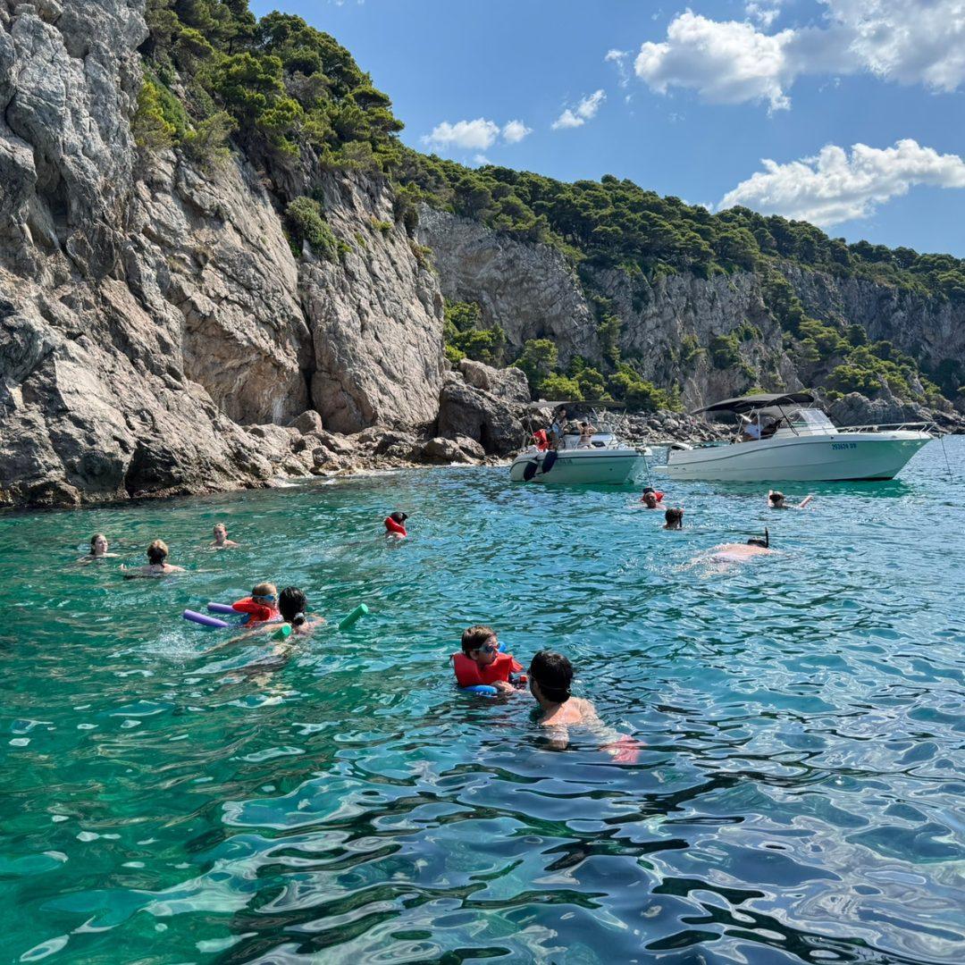 Group of swimmers and snorkelers exploring the clear green-tinted waters near the cliffs of Koločep (Kalamota) Island, with boats anchored nearby.