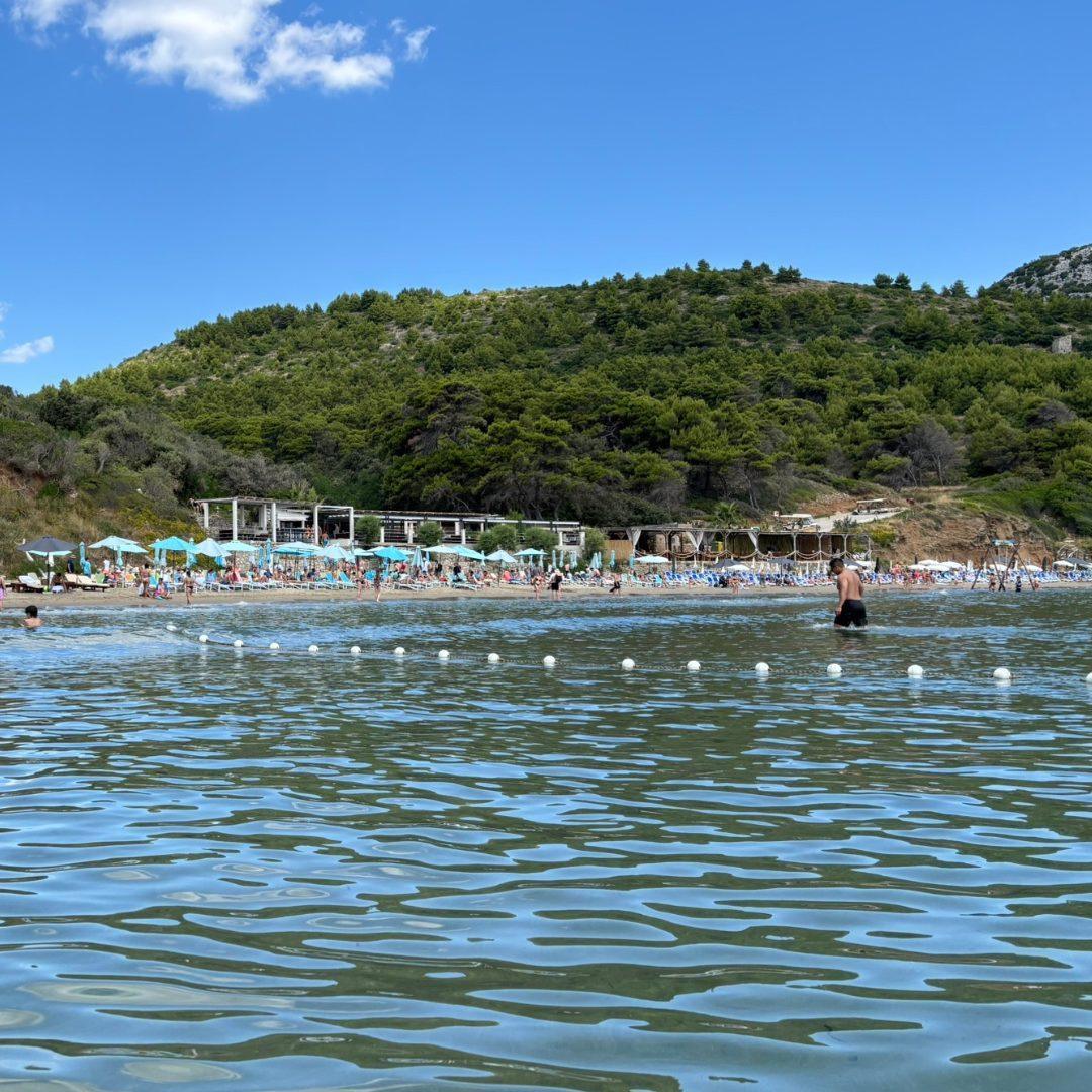  View of &Scaron;unj Beach on Lopud Island, with people swimming in shallow water and beach bars and loungers set against a green hillside.