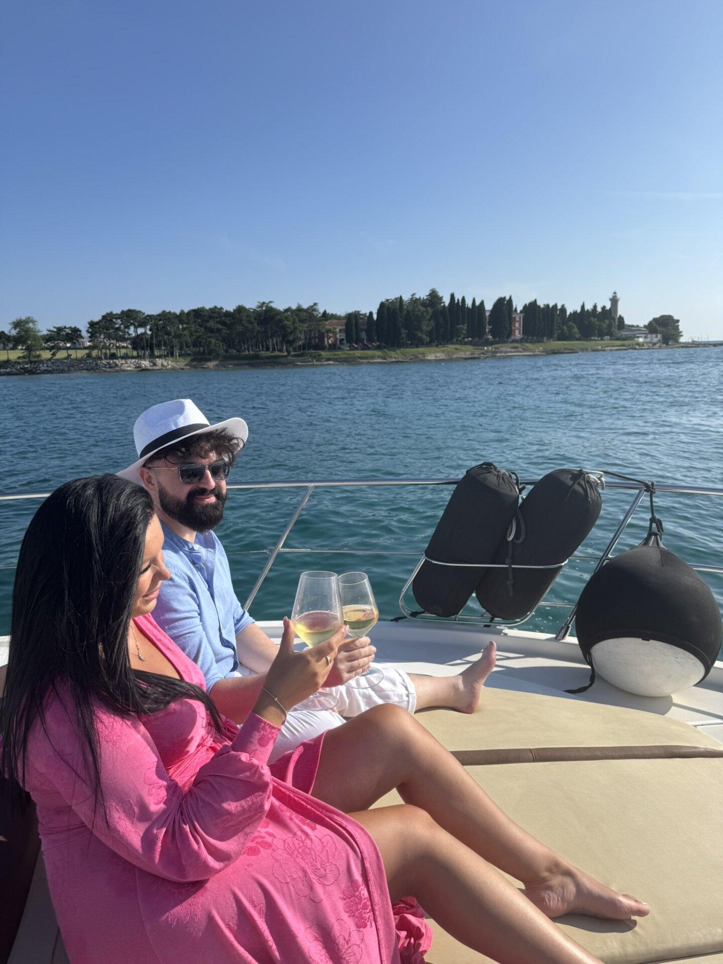 Ein Paar – sie in rosa Kleid, er mit weißem Hut – stößt auf einem Sonnendeck mit Weingläsern an