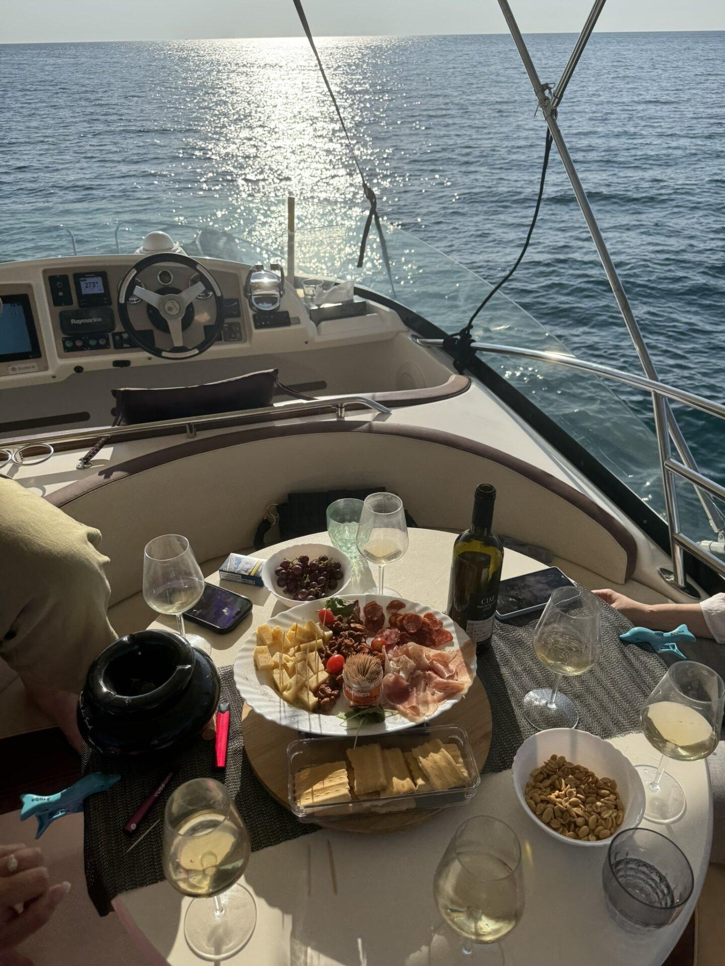 Aufschnitt, Wein und Snacks auf dem Cockpittisch mit dem Steuer und dem glitzernden Meer bei Sonnenuntergang.
