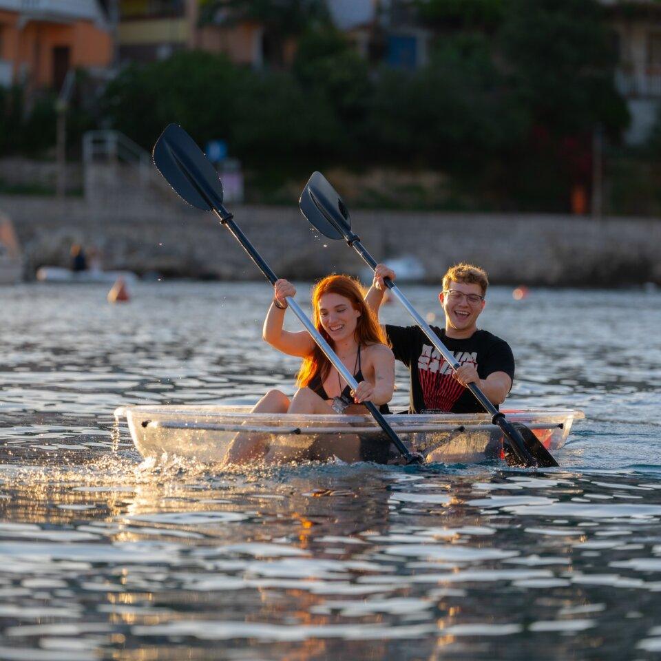 Two smiling kayakers paddling a transparent kayak on calm water near Rabac, Croatia, with village houses in the background