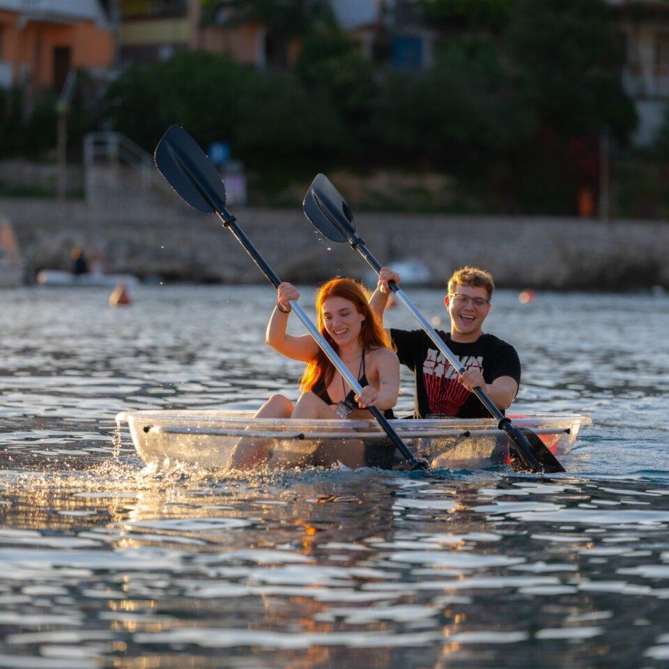 Zwei lächelnde Kajakfahrer erkunden in ihrem transparenten Kajak die ruhigen Gewässer vor Rabac, Kroatien, mit malerischen Dorfhäusern im Hintergrund.