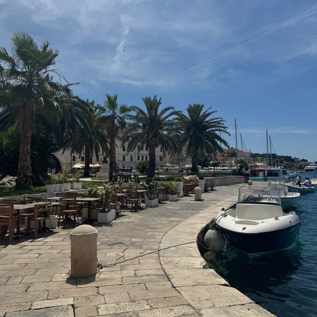 Palm-lined promenade in Hvar Town with outdoor cafés, boats moored along the waterfront, and a bright summer sky.
