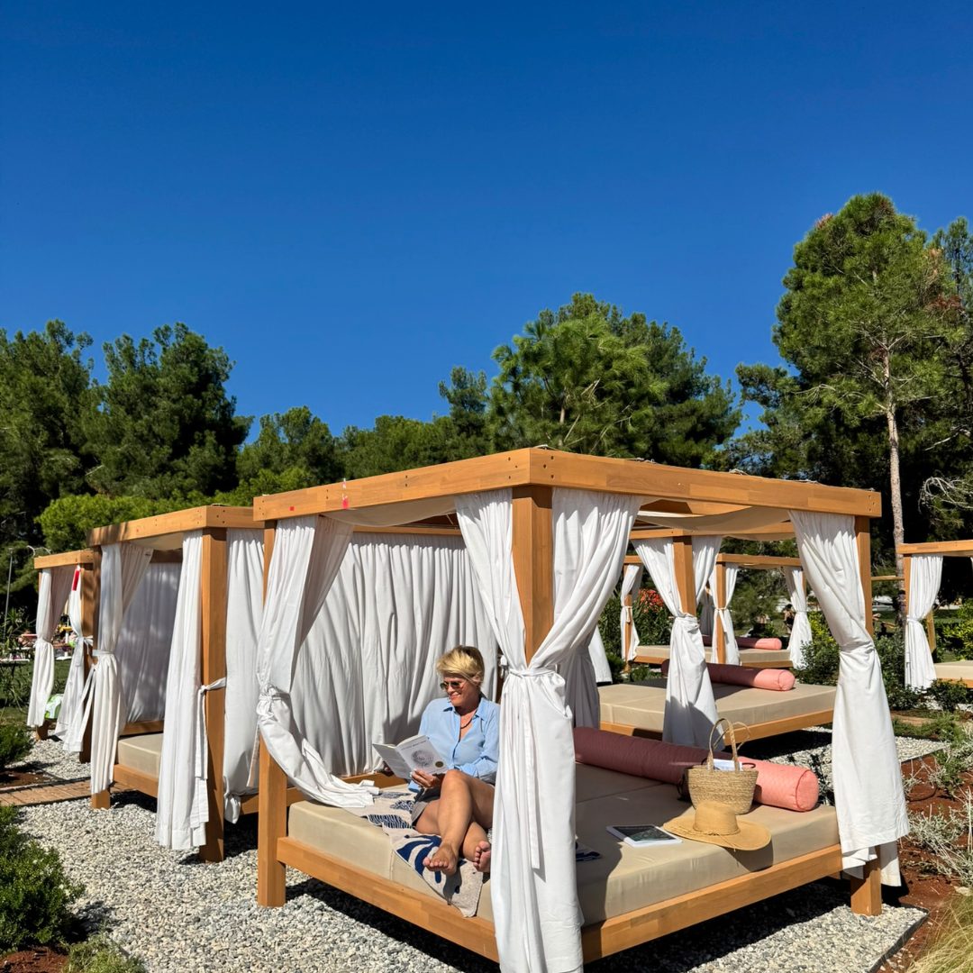 A person is relaxing on a shaded cabana bed with white curtains, reading a magazine.