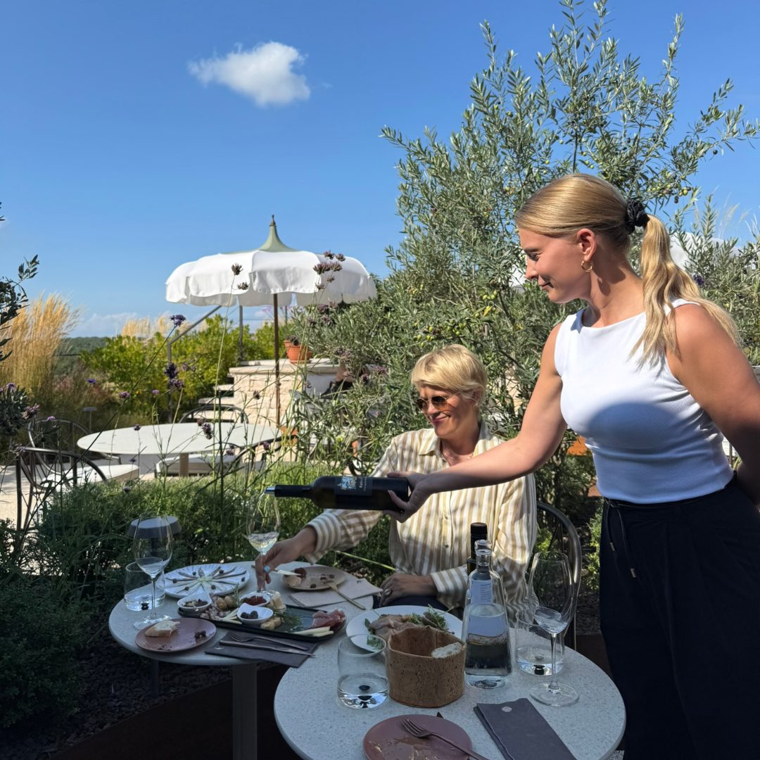 Woman pouring wine for a guest seated under a white umbrella at Radovan Wines terrace.