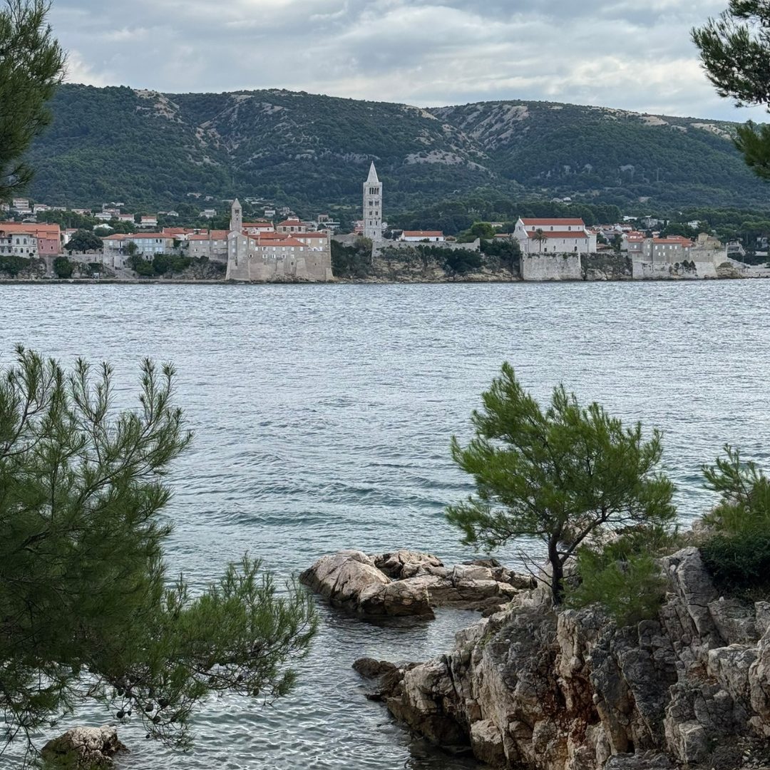 Scenic view of Rab’s old town with its historic bell towers, seen across the sea from a rocky pine-fringed coastline of the peninsula of Frkanj.