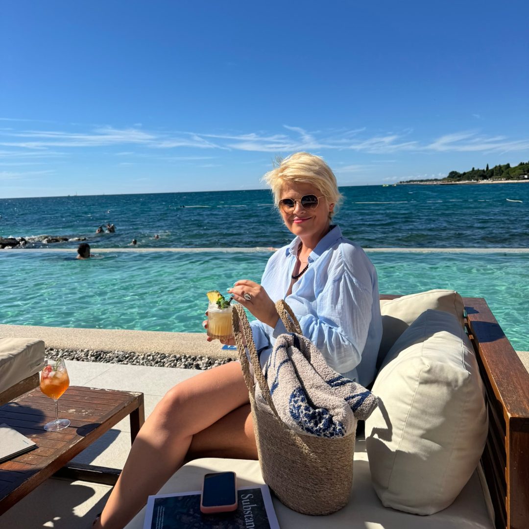 A person sitting on a lounge chair by the pool, holding a cocktail and smiling, with a beach bag, magazine, and phone on the side table.