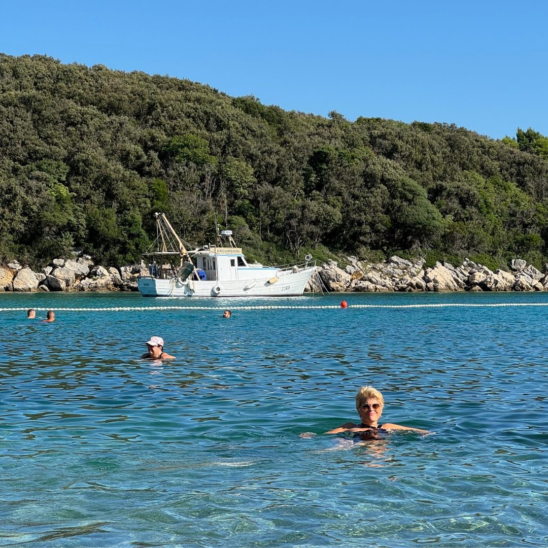 People swimming in the clear blue sea near Rab Island, with a white boat anchored by the forested coast.