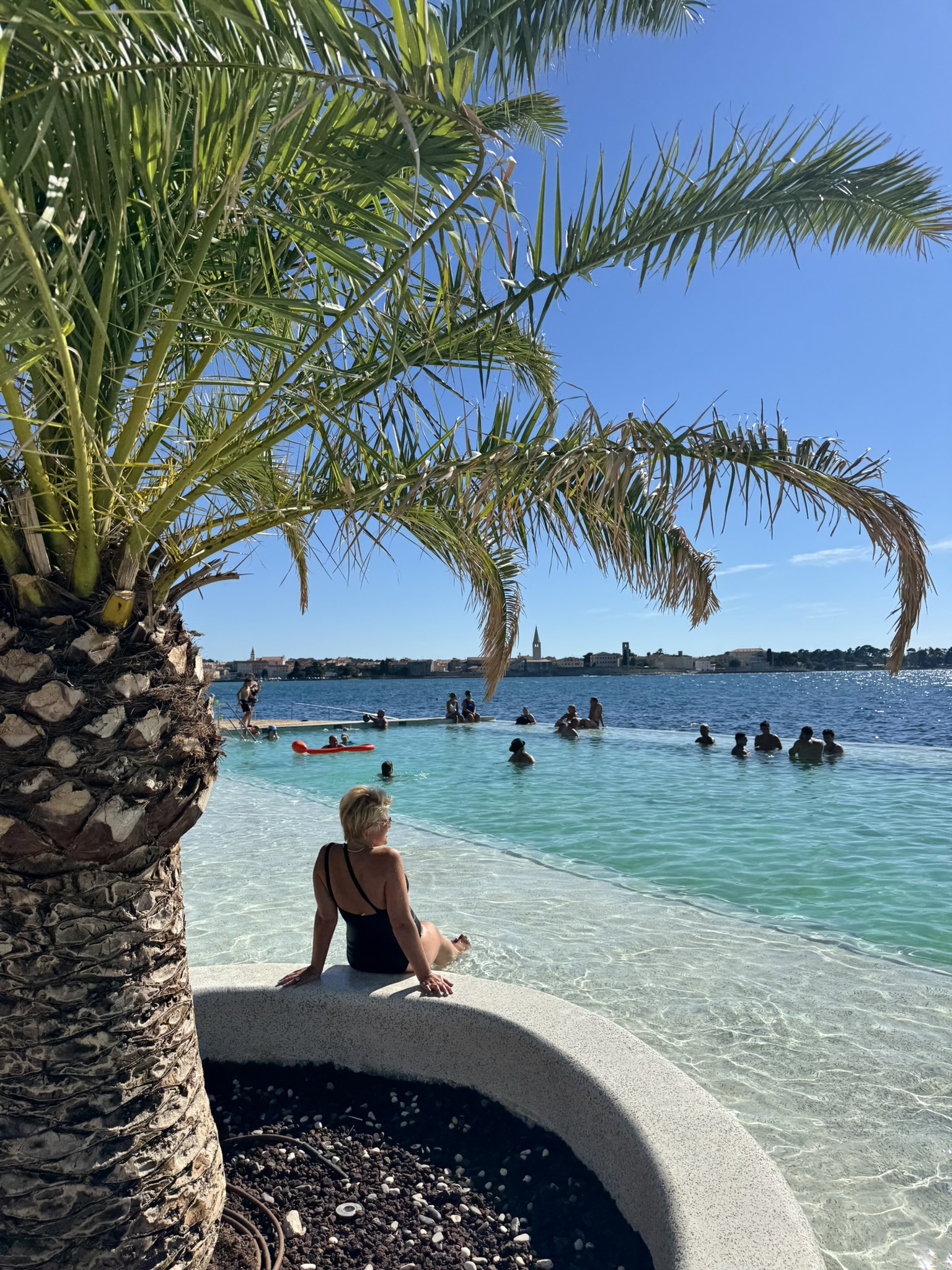 A woman enjoying the seaside infinity pool lined overlooking the calm blue Adriatic sea and Poreč old town in the distance.