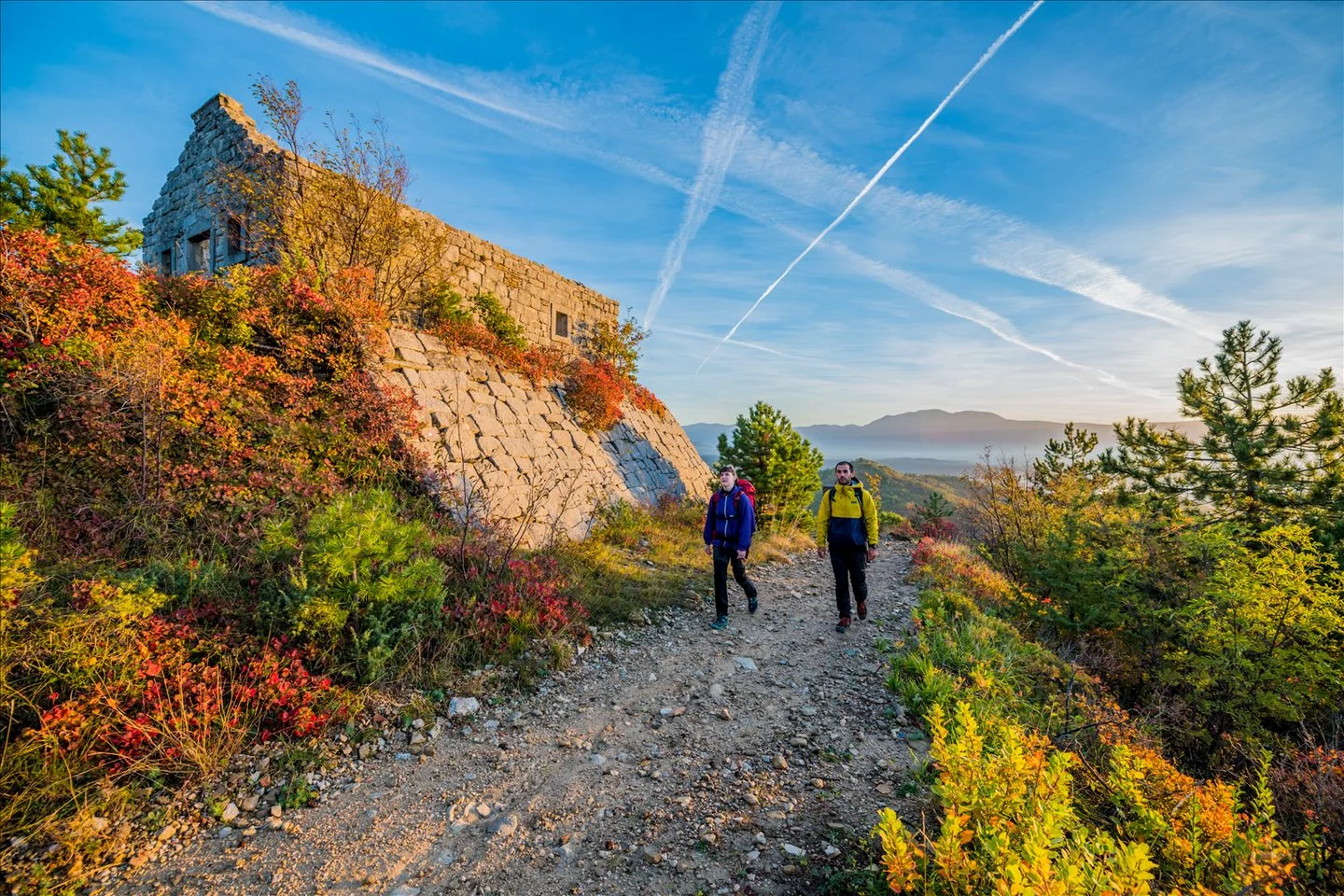 Zwei Wanderer gehen einen Pfad entlang, der an einem alten Steingebäude an einem Hang vorbeiführt, umgeben von herbstlicher Vegetation unter strahlend blauem Himmel.