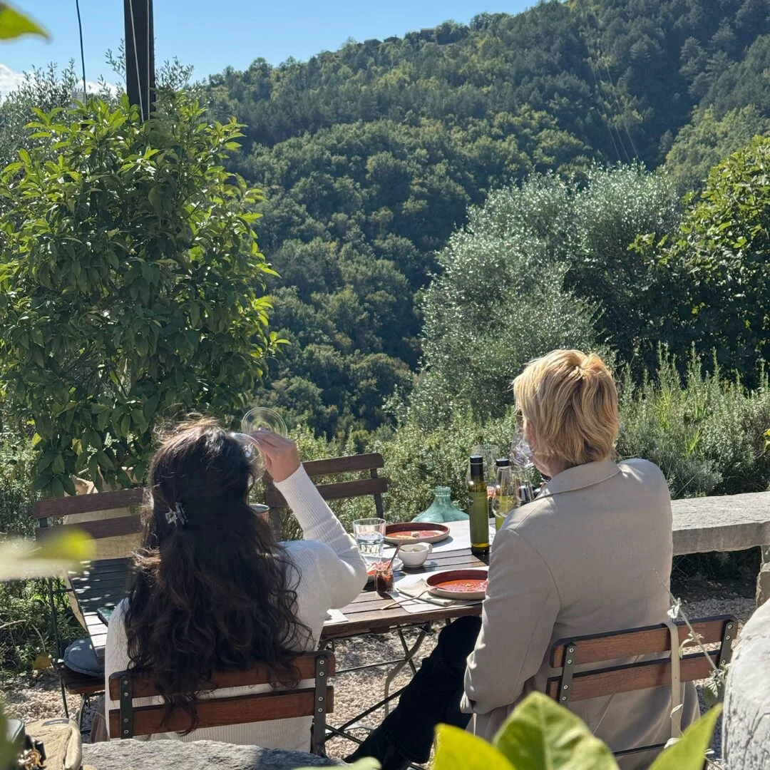 Zwei G&auml;ste genie&szlig;en eine Verkostung zu Mittag auf der Terrasse des Gutshofs Ip&scaron;a, mit einer Lichterkette und Ausblicken auf ein bewaldetes Tal in Istrien.