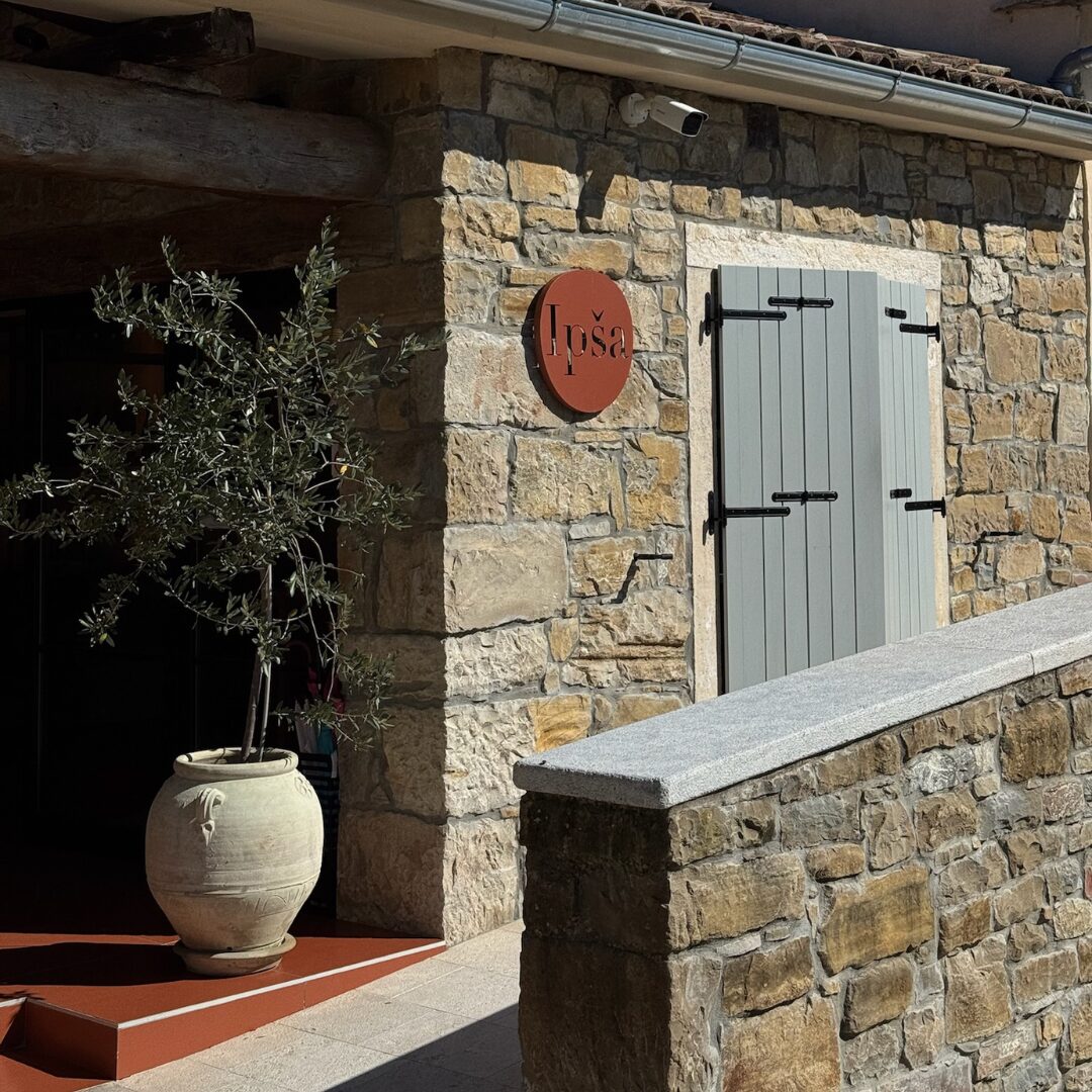 Stone façade of the Ipša estate with red round sign, blue shutters, and a potted olive tree.