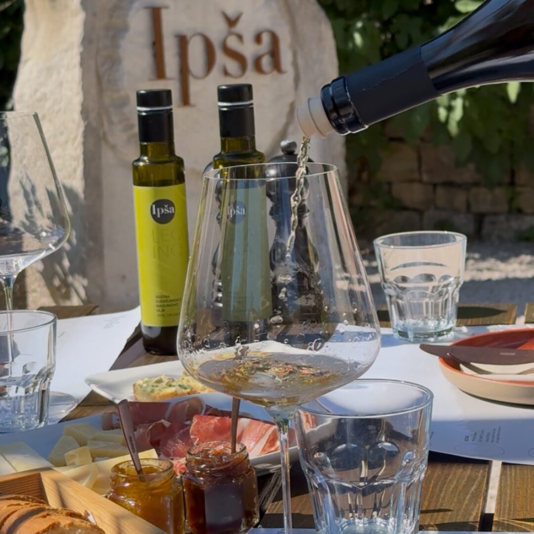 Close-up of a tasting table at Ipša with extra virgin olive oils, bread, cheeses, charcuterie, jams, and white wine being poured.