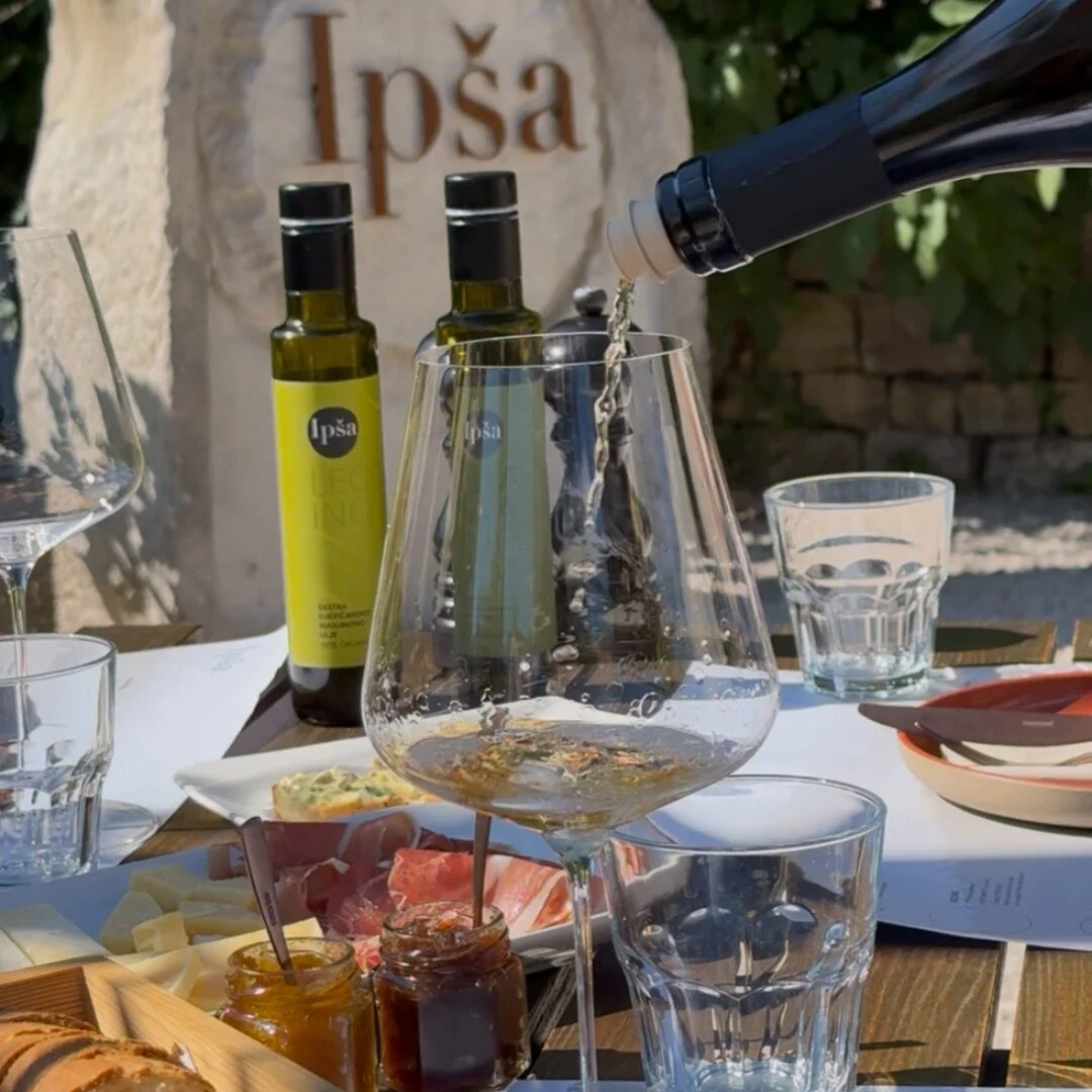 Nahaufnahme eines Verkostungstisches auf den Gutshof Ip&scaron;a, mit nativen Oliven&ouml;len extra, Brot, K&auml;se, Fleischwaren, Marmeladen und Wei&szlig;wein, der eingeschenkt wird. 