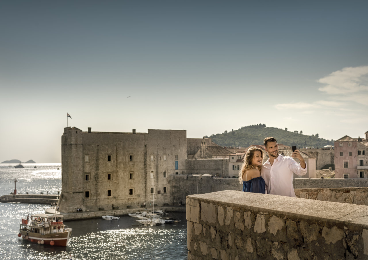 Couple taking a selfie on Dubrovnik’s ancient city walls with the Old Port and St. John Fortress shimmering in the background.
