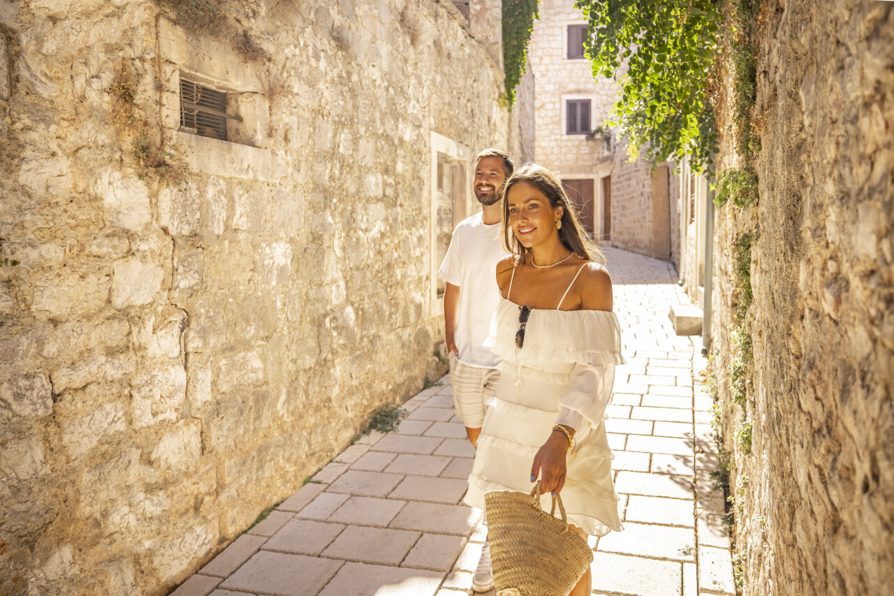 A smiling couple walks through a narrow stone alleyway in Stari Grad on a sunny day.