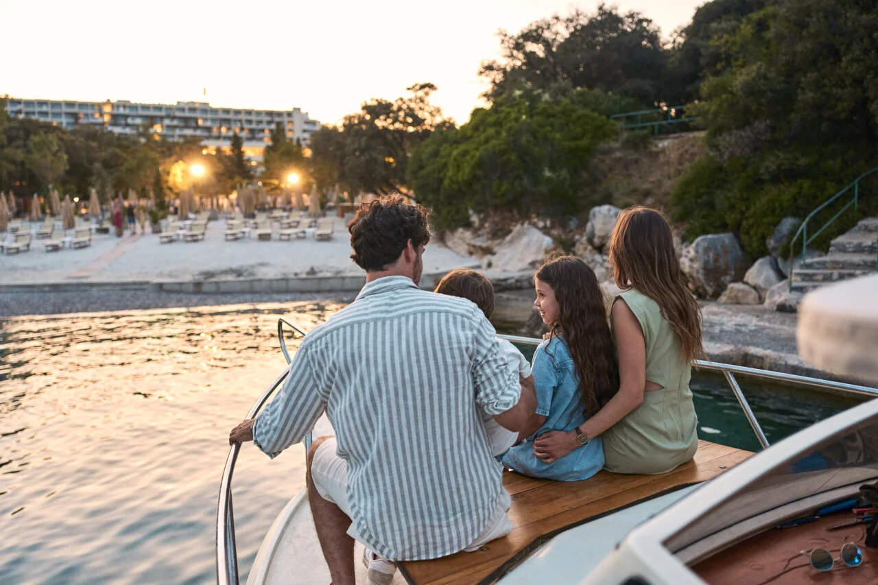 Eine vierk&ouml;pfige Familie am Bug eines Bootes bei Sonnenuntergang, auf einen Sandstrand mit Liegen zufahrend. 