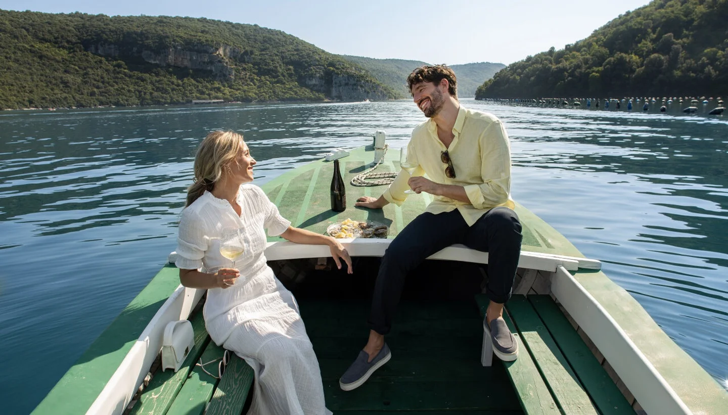 Ein Paar genießt eine exquisite Wein- und Meeresfrüchteverkostung auf einem kleinen Boot, umgeben von ruhigem, klarem Wasser und üppigen grünen Hügeln.