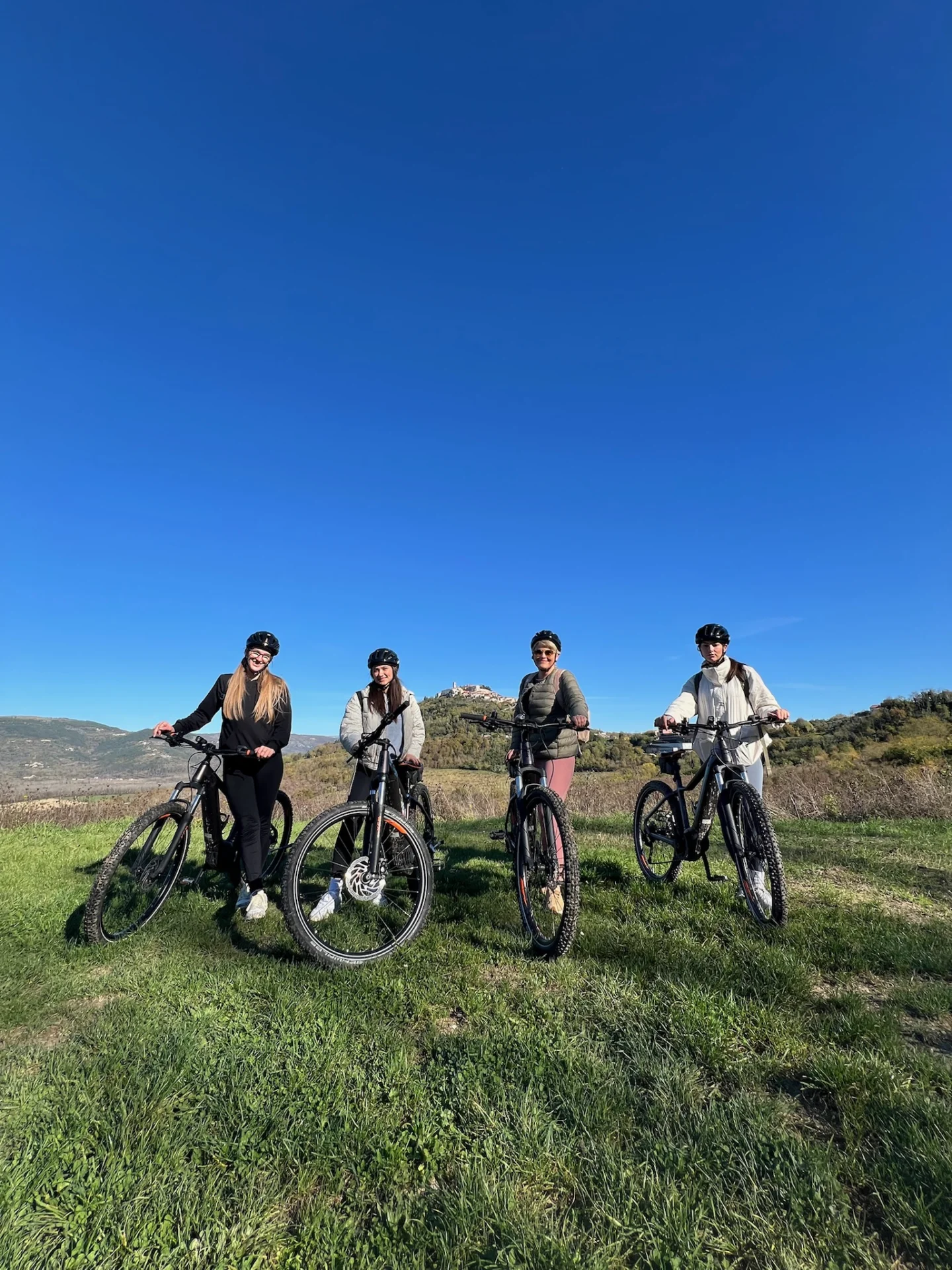 Vier Radfahrer mit Helmen und Mountainbikes auf einer gr&uuml;nen Wiese, die H&uuml;gelstadt Motovun unter strahlend blauem Himmel im Hintergrund.