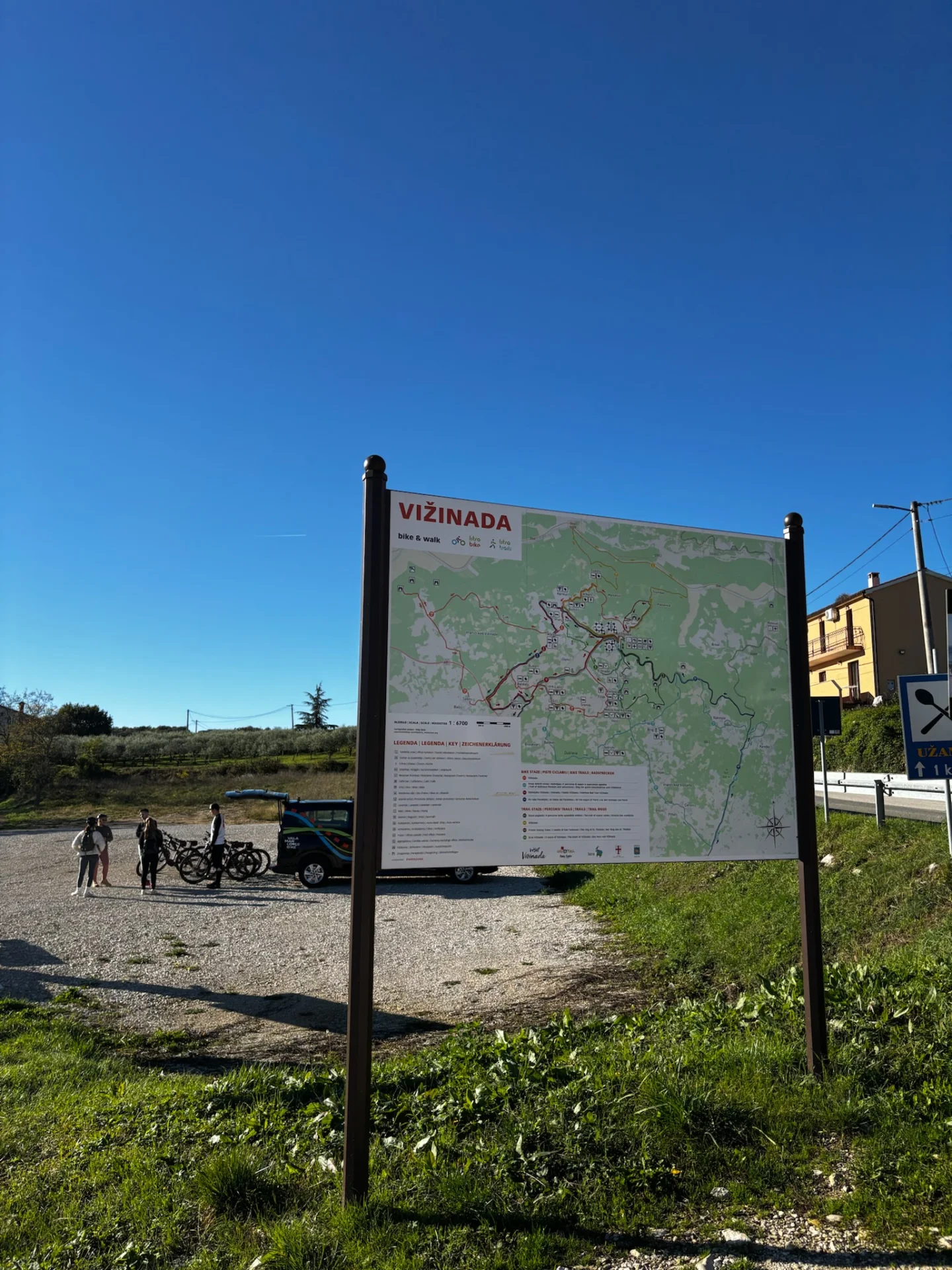 Unter strahlend blauem Himmel werden die Fahrr&auml;der vom Begleitfahrzeug auf dem Schotterplatz entladen, neben der Karte mit Rad- und Wanderwegen am Ausgangspunkt.