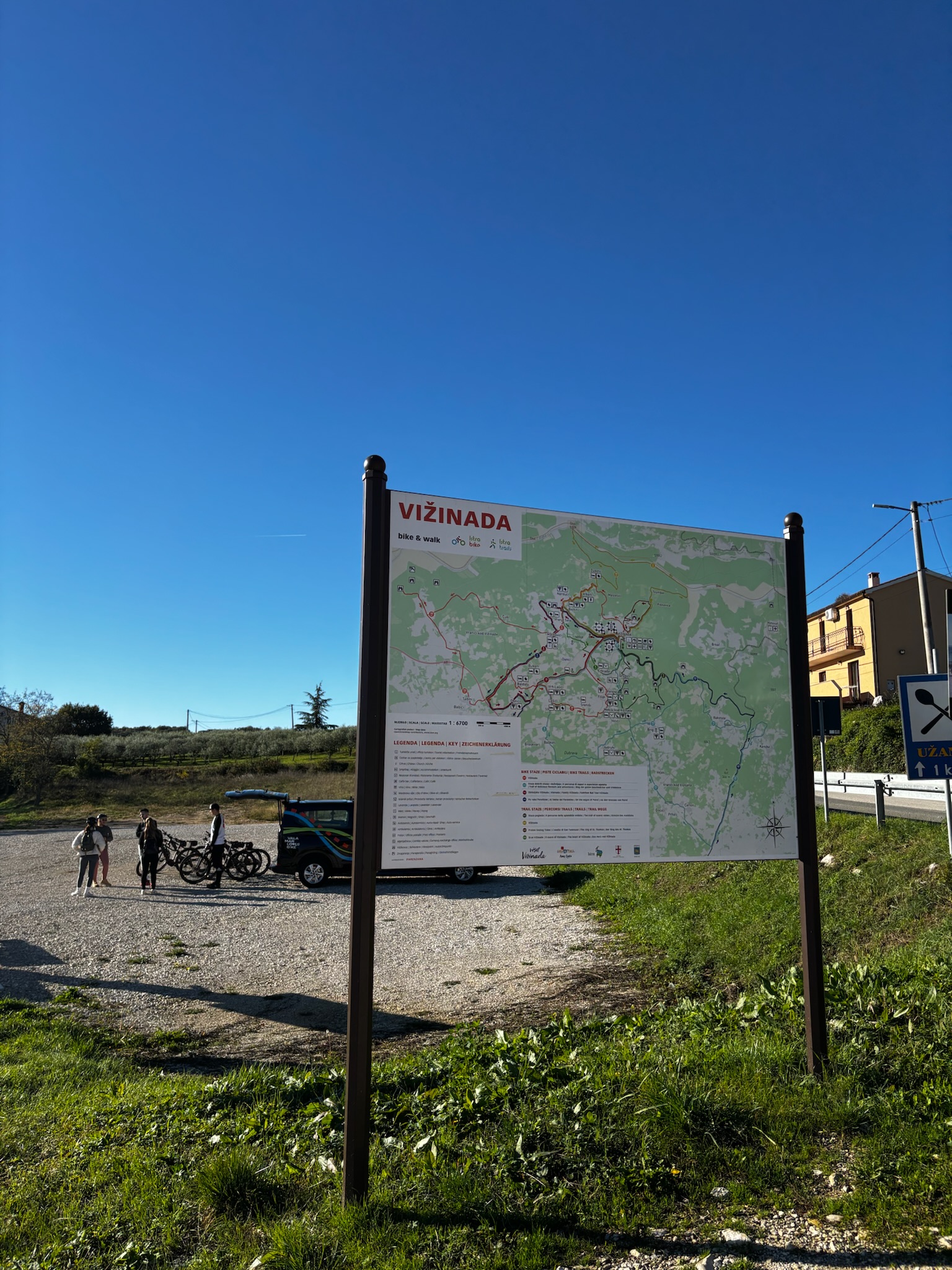 Vižinada trailhead map board for bike & walk routes, with a support van and cyclists unloading bikes in a gravel lot under a clear blue sky.