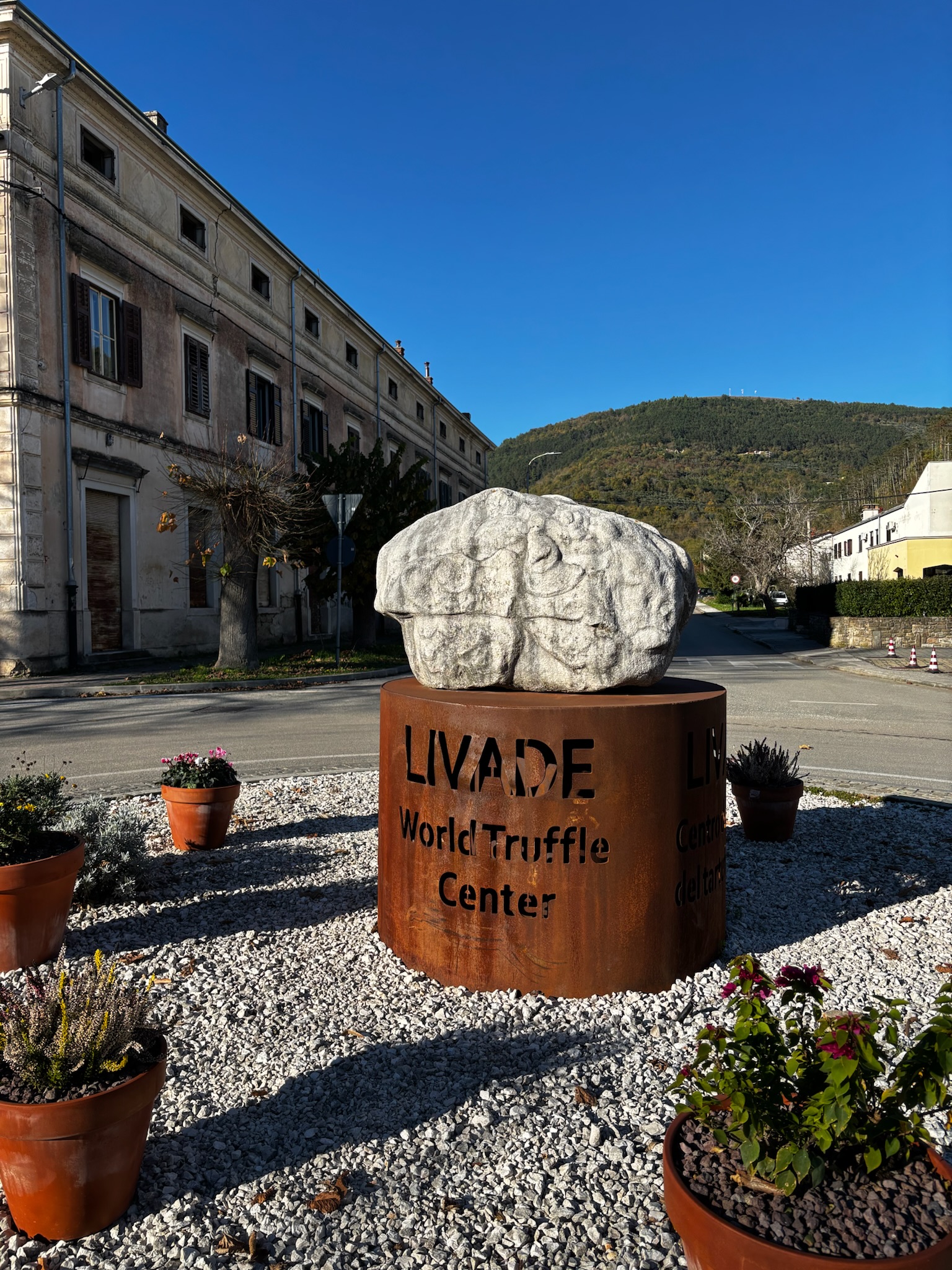 Stone truffle sculpture on a rusted steel pedestal reading &lsquo;LIVADE World Truffle Centre,&rsquo; in a village square with historic fa&ccedil;ades and a wooded hill under a clear blue sky.