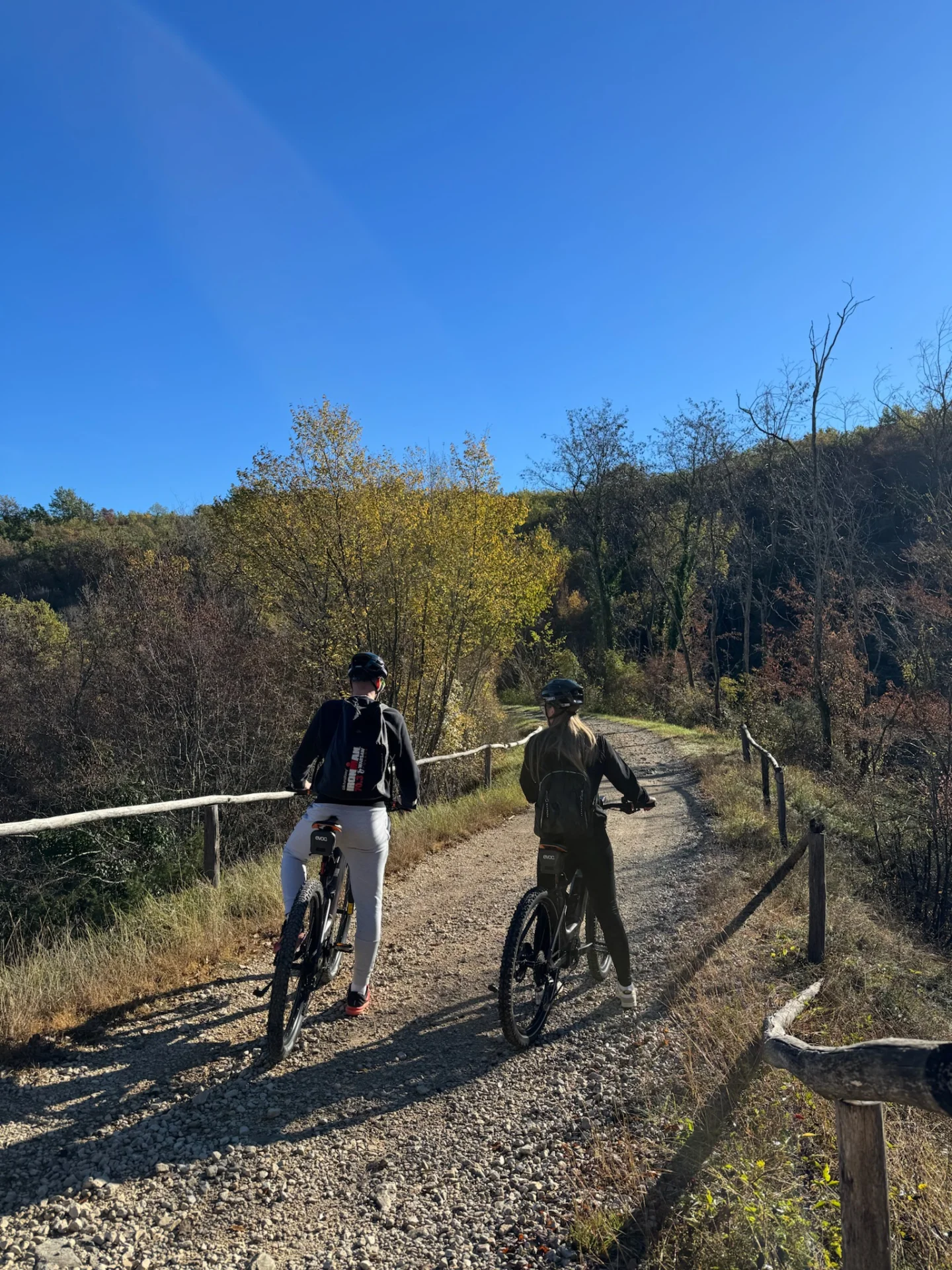 Zwei Radfahrer auf einer Schotterstrecke des Parenzana-Wegs, die sich unter strahlend blauem Himmel durch herbstliche W&auml;lder mit einem rustikalen Zaun schl&auml;ngelt. 