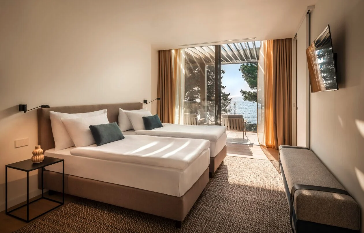Svijetla suvremena twin soba u Pical Hotelu s toplim neutralnim tonovima, staklenim stijenama od poda do stropa i privatnim balkonom s pogledom na more.