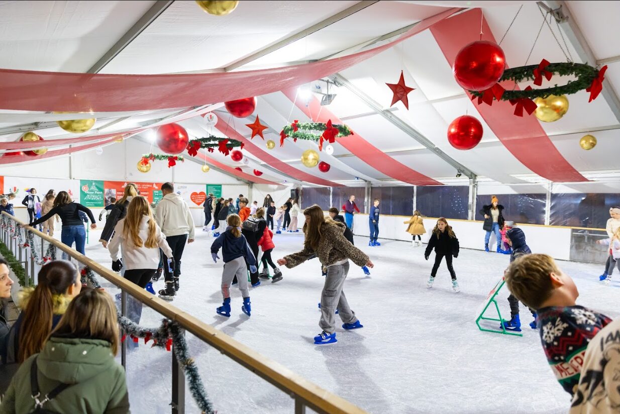 People of various ages are ice skating inside a festive indoor rink.