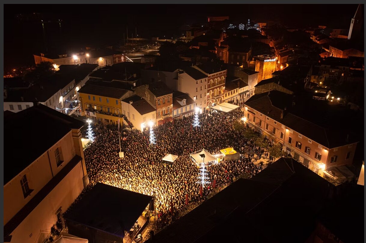 An aerial night view of a large crowd filling a town square for an outdoor holiday event, surrounded by historic buildings of Poreč and illuminated Christmas trees.