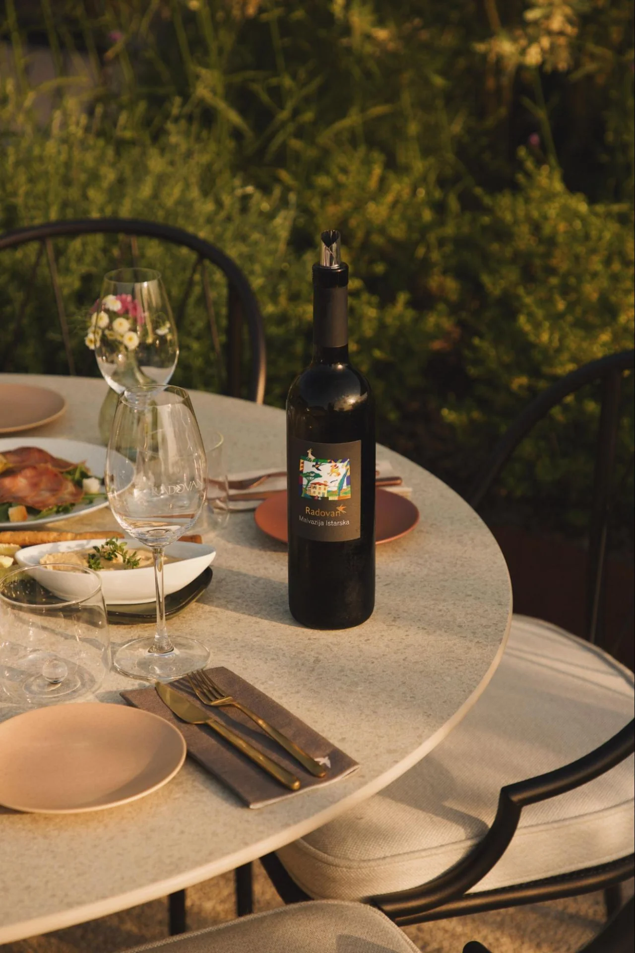 Ein gedeckter Tisch im Freien mit einer Flasche Rotwein, Gl&auml;sern und Tellern. 
