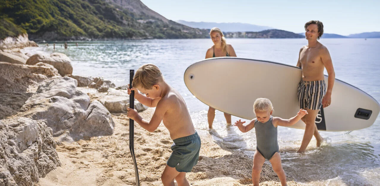 Eine Familie genießt einen Sommertag auf einem Kiesstrand in Istrien; Kinder spielen am Meeresufer und die Erwachsenen tragen ein SUP-Board