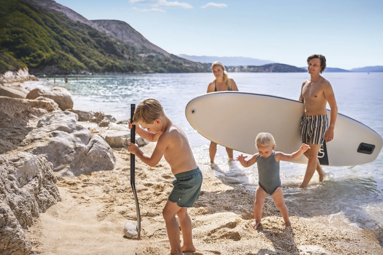 Obitelj uživa u ljetnom danu na &scaron;ljunčanoj plaži u Istri, djeca se igraju uz more, a odrasli nose SUP dasku.