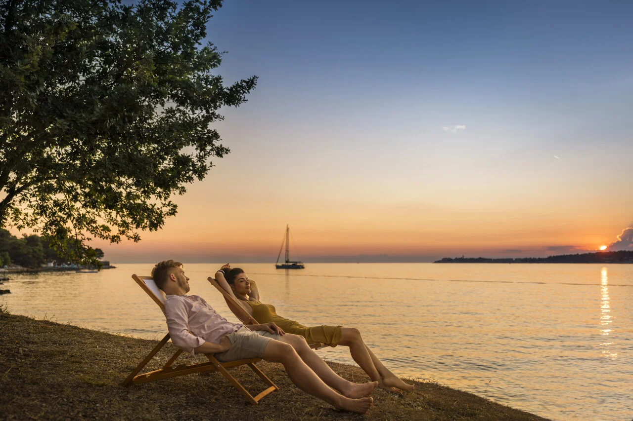 Ein Paar entspannt beim Sonnenuntergang auf Liegest&uuml;hlen am Adriatischen Meer, in der Ferne ist ein Segelboot zu sehen.
