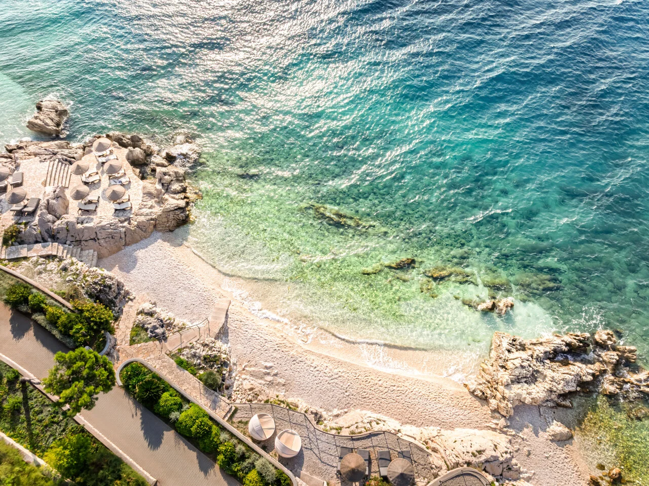 Pogled iz zraka na pješčanu plažu s tirkiznim morem, sunčanim kamenim terasama i obalnim putovima u Rapcu.