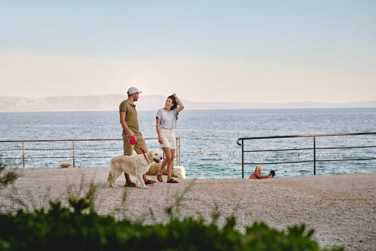 Ein Paar spaziert mit seinem Hund an einer Strandpromenade mit der Adria im Hintergrund. 