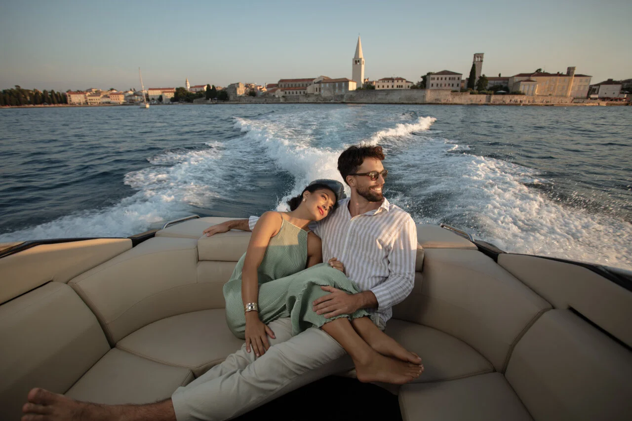 Ein Paar entspannt sich bei Sonnenuntergang auf einem Motorboot, im Hintergrund ist eine historische adriatische K&uuml;stenstadt zu sehen. 