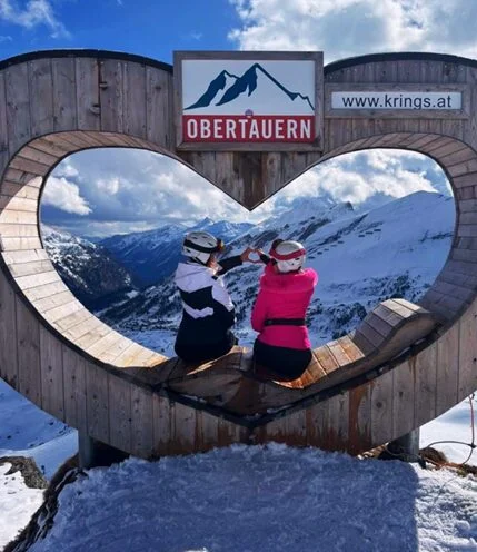 Two women posing on a large wooden heart, with a stunning alpine panorama in the background.
