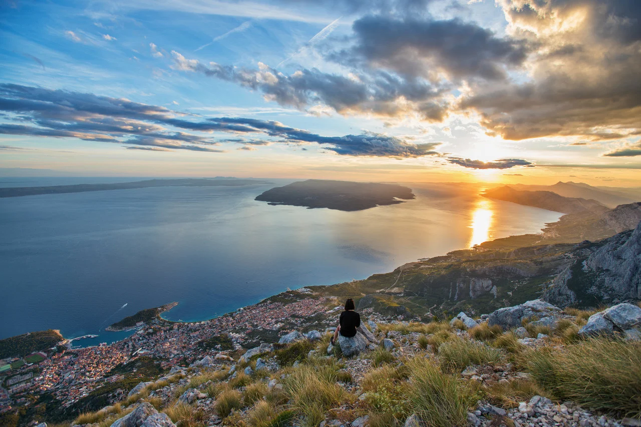 Djevojka sjedi na stjenovitoj planinskoj padini s pogledom na Jadran i Makarsku u suton.
