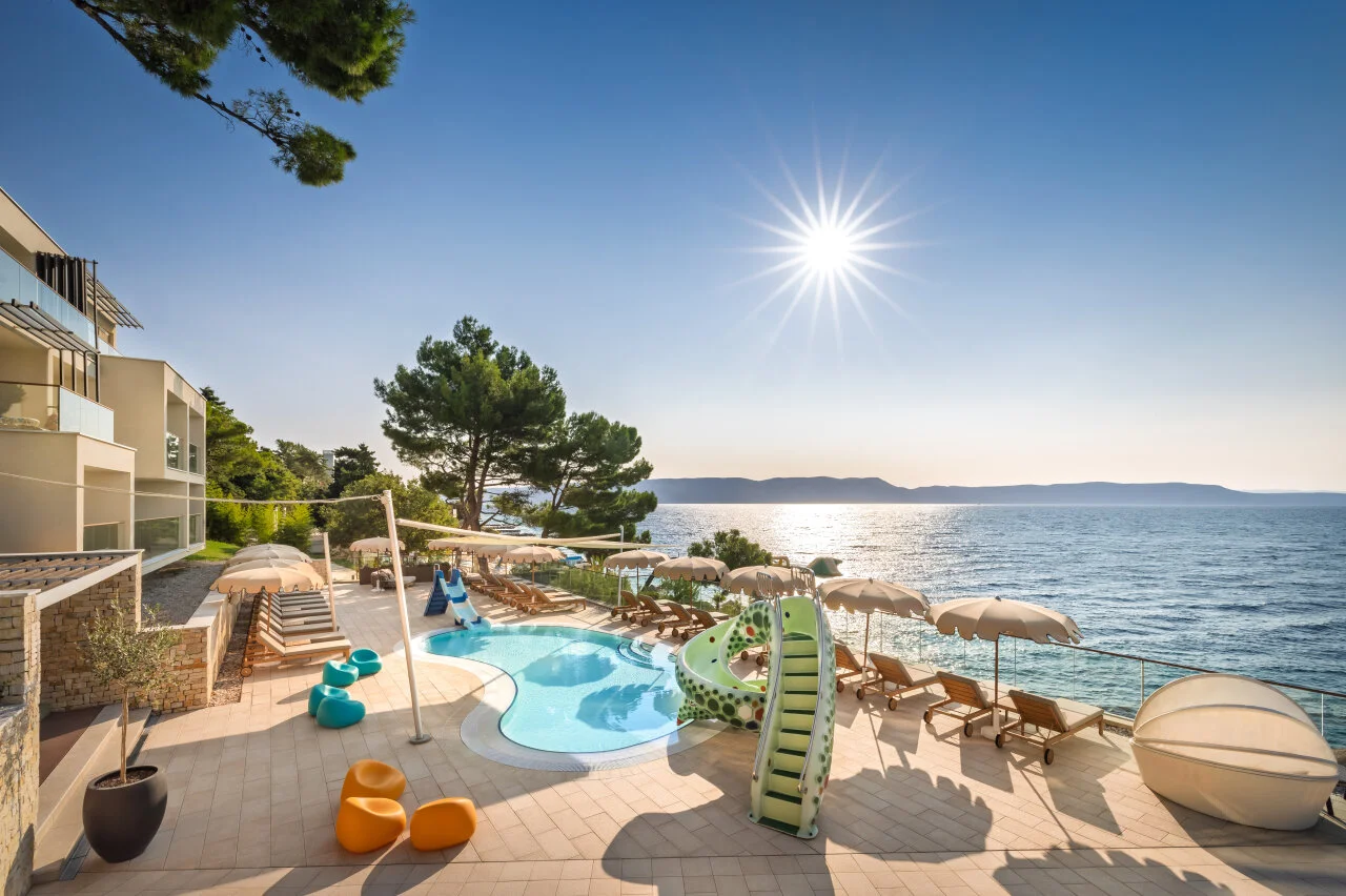 A seaside hotel pool area at Girandella Resort in Rabac with a children’s slide, sun loungers, and umbrellas overlooking the sparkling Adriatic Sea under a bright blue sky
